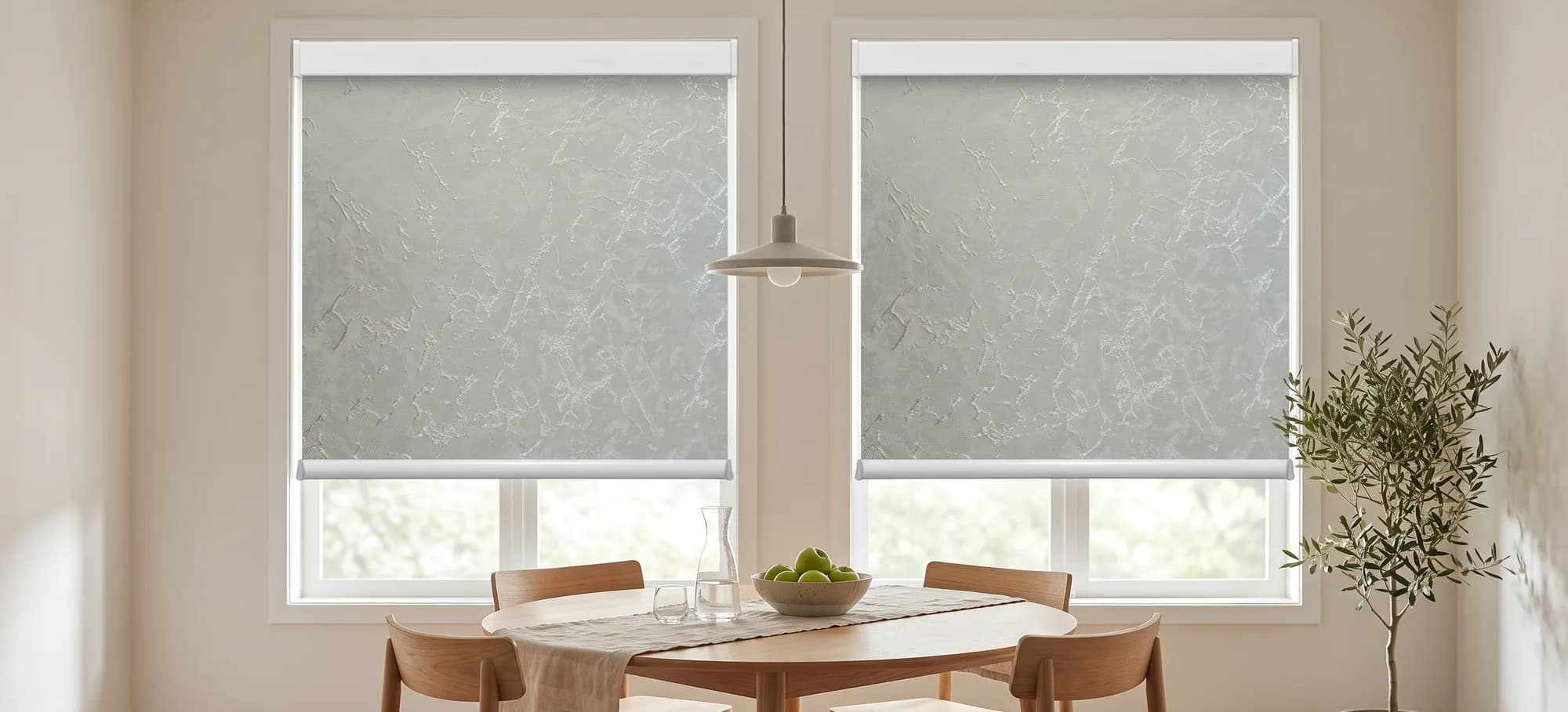 Pale sage roller shades with a soft marble pattern on two windows above a round oak dining table.